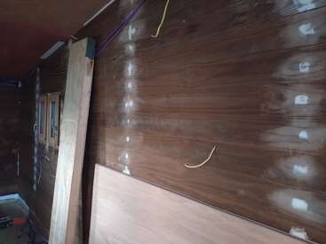 Das Bild zeigt einen teilweise renovierten Holzraum mit ausgebesserten Wänden, freiliegenden Kabeln, an der Wand angelehnten Baumaterialien und einem kleinen Fenster auf der linken Seite.