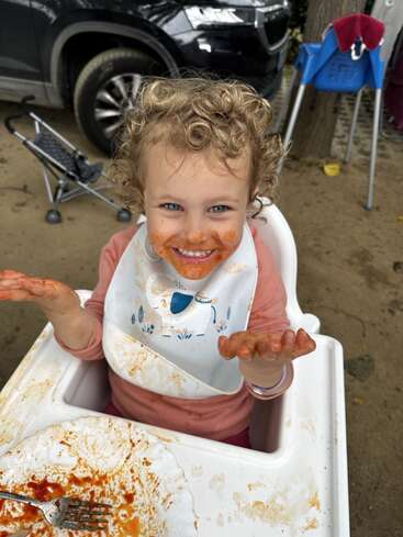 A young child with curly blonde hair sits in a high chair, covered in food, with a bib on and a messy tray in front of them.