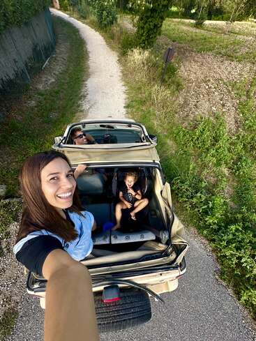 The image shows a woman taking a selfie while riding in the back of a convertible car with two people and a child in the front seat on a gravel path.
