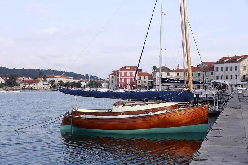 La imagen representa un velero atracado en un puerto deportivo, con el casco de madera y el interior blanco, y una lona azul cubriendo la cubierta, sobre un fondo de edificios y un cielo nublado.