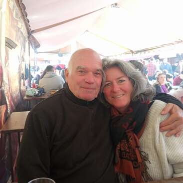 Un couple âgé, tout sourire, est assis côte à côte sur un marché en plein air très animé ou une foire, profitant de la compagnie l'un de l'autre. L'ambiance autour d'eux est animée et chaleureuse.