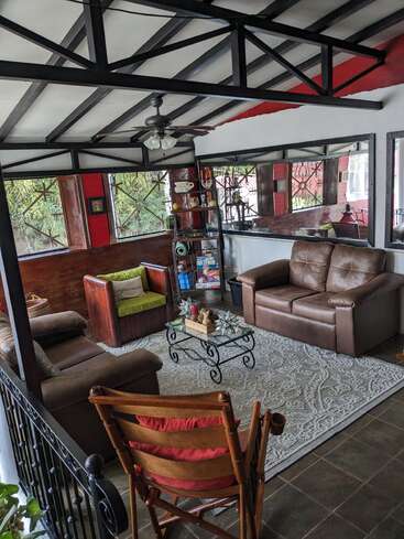 Essa aconchegante sala de sol tem sofás de couro, uma poltrona de madeira, mesa de centro de vidro, estante, tapete grande, ventilador de teto, almofadas decorativas e muita luz natural.