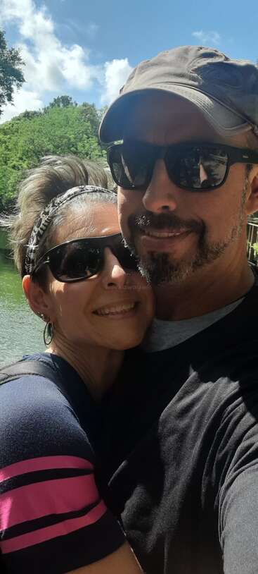 Um casal feliz posa para uma selfie ao ar livre em um dia ensolarado, ambos usando óculos escuros e sorrindo, com árvores verdes, céu azul e água atrás deles.