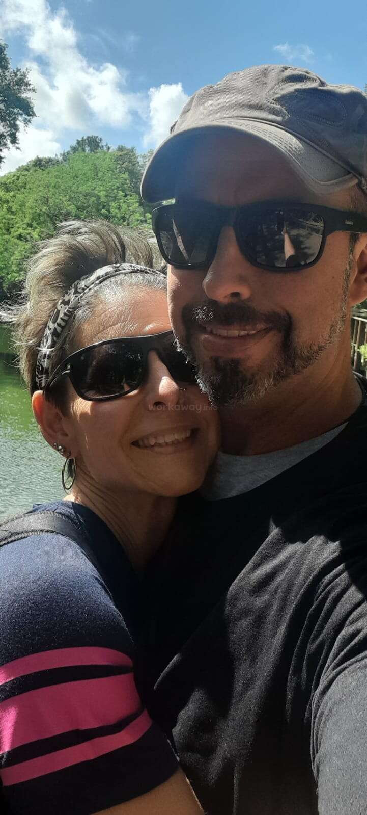 Um casal feliz posa para uma selfie ao ar livre em um dia ensolarado, ambos usando óculos escuros e sorrindo, com árvores verdes, céu azul e água atrás deles.