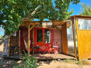 A colorful, cozy wooden cabin with a decorated red door sits under a leafy tree. A small porch features red chairs, a table, and natural sunlight.