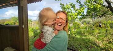 A woman with glasses joyfully holds a smiling toddler outdoors. Surrounded by lush greenery, their laughter and happiness create a warm, loving, and natural atmosphere.