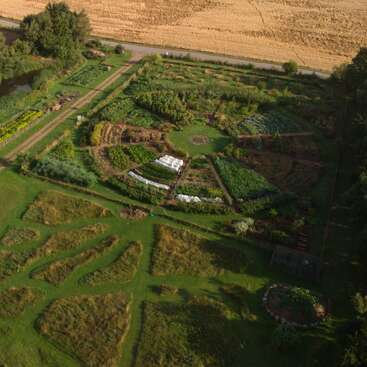 Essa foto aérea mostra uma grande horta circular dividida em seções, cercada por caminhos gramados, campos, árvores e uma estrada por perto.