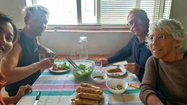 Four people sit around a table with a colorful checkered tablecloth, enjoying a meal of spring rolls, salad, and small dishes, smiling and sharing conversation.