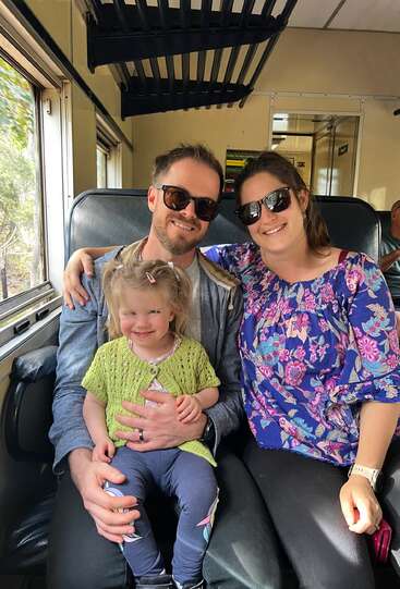 The image shows a family of three sitting on a train, with the man and woman embracing their young daughter, who is wearing a yellow cardigan.