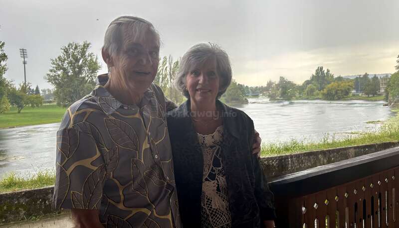 Um casal de idosos está junto, sorrindo em uma varanda com vista para um rio tranquilo, com vegetação exuberante e árvores ao fundo, sob um céu nublado.