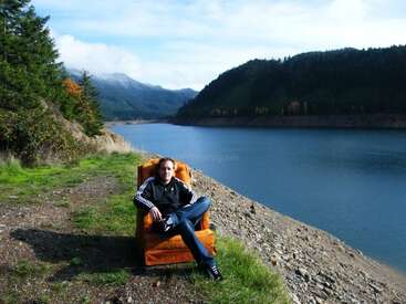 Un hombre con chaqueta negra se sienta cómodamente en un sillón naranja junto a un lago, rodeado de montañas, árboles y un cielo claro y brillante.