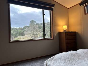 A cozy bedroom features a large window overlooking greenery, a wooden dresser with a glowing lamp, beige walls, grey carpet, and a neatly made bed.