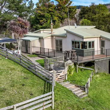 This image shows a modern rural house with light-colored siding, surrounded by lush greenery, wooden fences, a deck, sloped yard, and forested hills in the background.