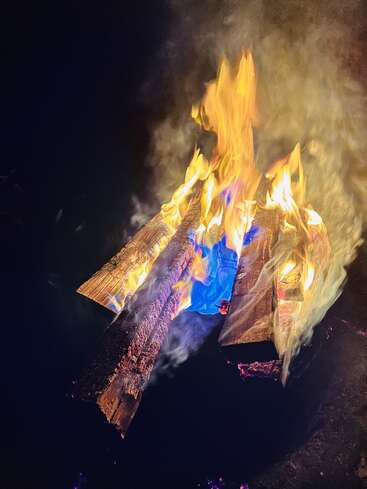 Tres troncos de madera arden en un entorno oscuro. Las llamas parpadean en vibrantes tonos amarillos, naranjas y azules, y el humo se arremolina en el aire nocturno.