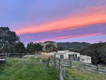 Un sereno paisaje rural al atardecer, con una granja, pastos verdes, árboles, vallas de madera, colinas al fondo y vibrantes nubes rosas y moradas en el cielo.