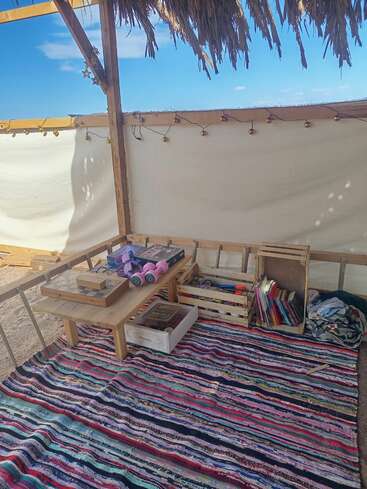 This cozy outdoor nook features a colorful woven rug, wooden table with games and dumbbells, crates of toys, books, string lights, and a thatched roof above.