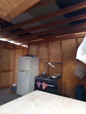 This image shows a simple wooden kitchen with a refrigerator, gas stove, and cooking pots. The roof is unfinished, and a gas cylinder is visible.