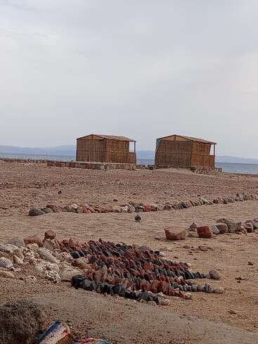 Two small wooden huts stand on a rocky shore, overlooking the sea, surrounded by lines of colorful stones and pebbles under a cloudy sky. Peaceful scene.