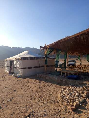 A traditional tent and shaded seating area stand in a desert landscape, surrounded by mountains. The sun is shining brightly, casting long early morning shadows.