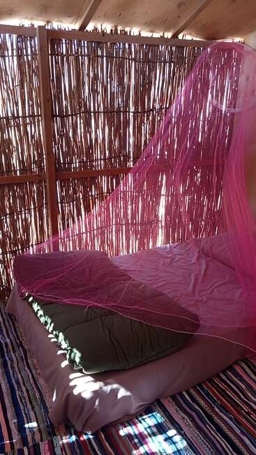 This image shows a rustic sleeping area with a bed covered in a pink mosquito net, set against bamboo walls and a colorful striped rug on the floor.