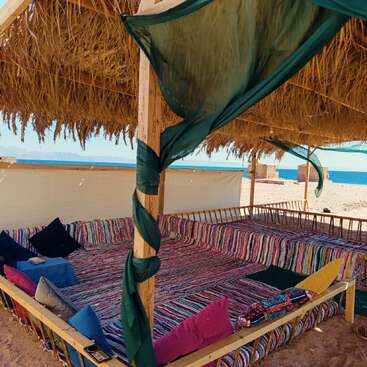 A cozy beach hut with a thatched roof, colorful striped seating, and assorted cushions overlooks sandy shores and a bright blue sea under clear skies.