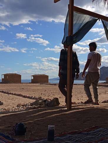 Two men stand under a shelter, looking at wooden huts by the sea. The sky is filled with clouds. Tea glasses and a teapot are nearby.