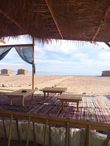 This image shows a shaded, rustic seating area with wooden tables on colorful rugs, overlooking a sandy beach with huts and a tranquil sea view.