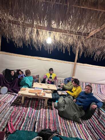 Six people relax on patterned rugs under a thatched roof, sharing food and drinks around low tables, wrapped in blankets and sleeping bags under a hanging light.
