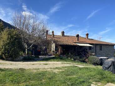 A rustic stone house with a red-tiled roof sits under a blue sky, surrounded by greenery, leafless trees, and scattered garden items in the yard.