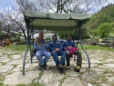Tres hombres están sentados juntos en un columpio verde al aire libre con un toldo, sonrientes y relajados. Detrás de ellos hay árboles, suelo de piedra y un jardín frondoso y verde.