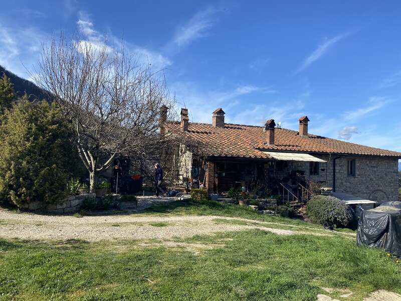 A rustic stone house with a red-tiled roof sits under a blue sky, surrounded by greenery, leafless trees, and scattered garden items in the yard.