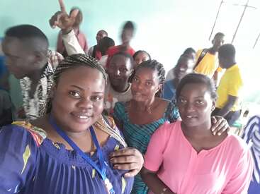 A group of people gather indoors, smiling and posing for a selfie. The atmosphere is lively, with various individuals in colorful clothing, expressing happiness and camaraderie.