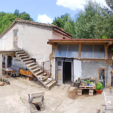 This image shows a rustic, weathered countryside house with outdoor stairs, potted plants, simple furniture, and a lush green background. Sunlight and shadows create a peaceful atmosphere.