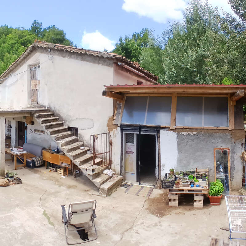Cette image montre une maison de campagne rustique, vieillie par le temps, avec des escaliers extérieurs, des plantes en pot, des meubles simples et un arrière-plan verdoyant. La lumière du soleil et les ombres créent une atmosphère paisible.