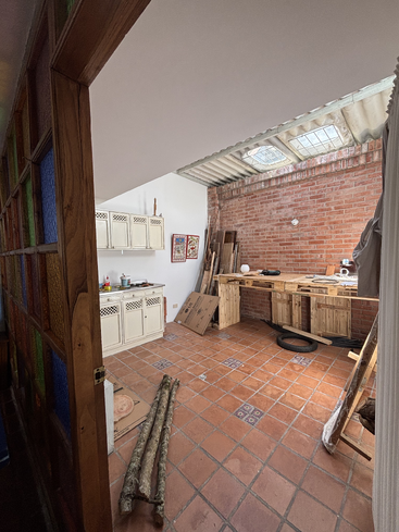 This room has terracotta floor tiles, wooden workbenches, exposed brick walls, skylight roofing, white cabinets, colorful glass-paneled door, and some construction materials scattered around.