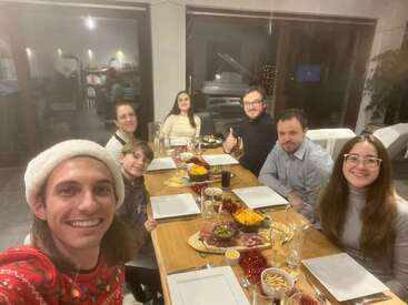 Sete pessoas estão reunidas em torno de uma mesa de jantar festiva, sorrindo para uma selfie. Há petiscos, pratos vazios e bebidas, criando uma atmosfera calorosa e alegre de celebração.