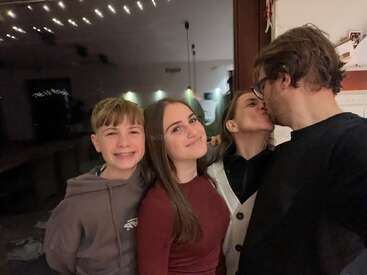 Quatro pessoas posam juntas dentro de casa, sorrindo calorosamente. Dois adultos se beijam carinhosamente, enquanto dois jovens ficam próximos, parecendo felizes e relaxados. Um momento familiar aconchegante capturado.