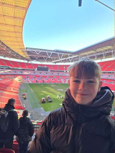 Um menino sorridente com uma jaqueta preta está dentro do Estádio de Wembley. Os assentos do estádio são vermelhos e estão vazios, com veículos de manutenção no gramado verde abaixo.