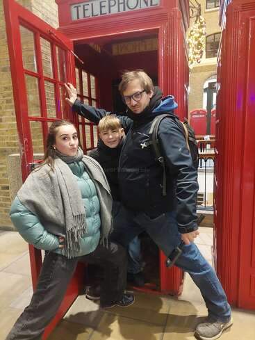 Três pessoas posam de forma divertida na frente de uma cabine telefônica vermelha clássica britânica. Elas parecem alegres e relaxadas, vestidas com roupas quentes para o tempo frio, curtindo o passeio.