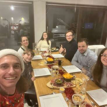 Seven people are gathered around a festive dinner table, smiling for a selfie. There are snacks, empty plates, and drinks, creating a warm, joyful celebration atmosphere.