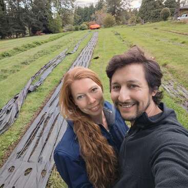 Un couple souriant prend un selfie en plein air dans une ferme herbeuse dont les rangées sont recouvertes de plastique noir, entourée d'arbres et de verdure sous un ciel nuageux.