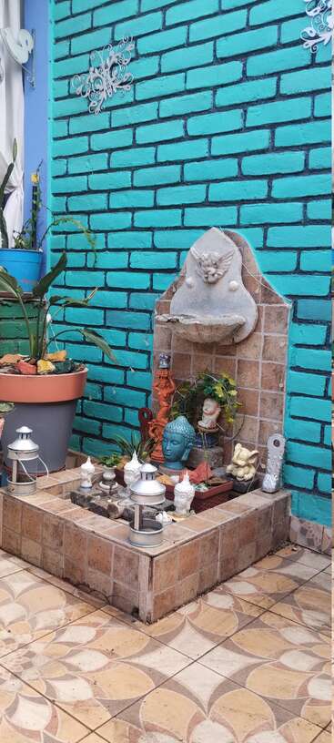 Ein kleiner, gefliester Brunnen steht vor einer türkisfarbenen Backsteinmauer, umgeben von Buddha-Köpfen, Laternen, Pflanzen, Statuen und dekorativen Elementen, die eine friedliche Gartenecke bilden.