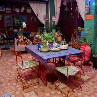 The image depicts a cozy outdoor patio with a blue table, red chairs, and colorful decorations, surrounded by plants and string lights.