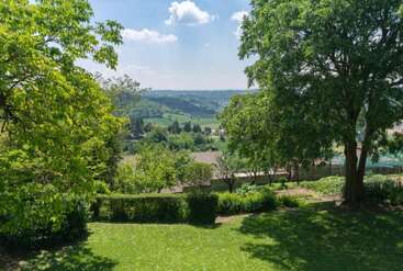 This image displays a lush green garden with large trees, neatly trimmed bushes, and vibrant grass, overlooking a scenic valley and distant hills under a bright sky.