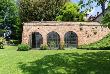 This image shows a brick building with three large arched windows, surrounded by lush green grass, bushes, and tall trees under a bright sky.