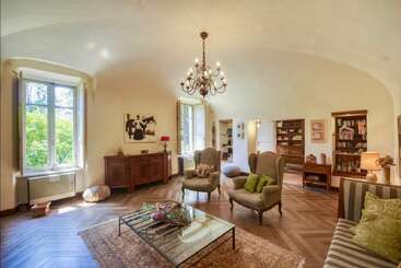 This elegant living room features high ceilings, large windows, vintage armchairs, a glass coffee table, a chandelier, hardwood floors, bookshelves, and tasteful, cozy decor.