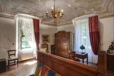 This image shows an elegant, vintage bedroom with ornate ceiling, chandelier, large wooden bed, antique furniture, deep red curtains, and sunlight streaming through tall windows.