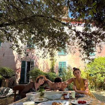 Three people sit at an outdoor table covered with food, under tree shade, in front of a rustic house with green shutters, enjoying a relaxed summer meal.