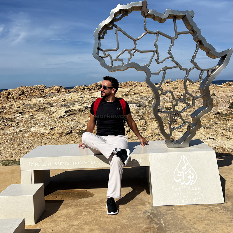 A man with sunglasses and a red backpack sits on a concrete bench near a modern metal Africa sculpture, with rocky ground and blue sky in the background.
