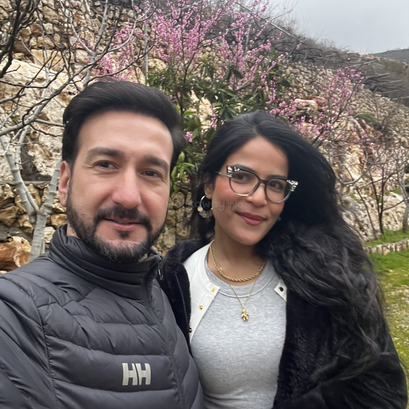 Un hombre y una mujer sonrientes posan para hacerse un selfi al aire libre, de pie frente a unos árboles en flor con flores rosas y un terreno rocoso, disfrutando de un día nublado.
